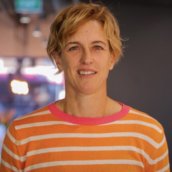 Person with short blonde hair wearing an orange and white striped sweater with a pink collar, standing indoors.