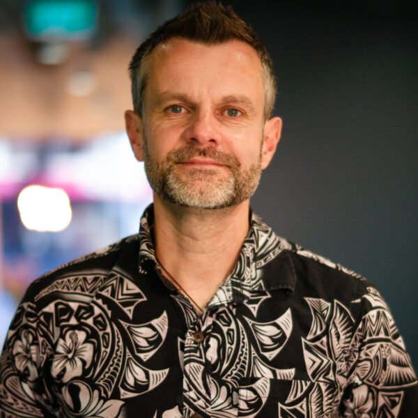 Man with short hair and beard wearing a black and white patterned shirt, standing indoors with blurred background.