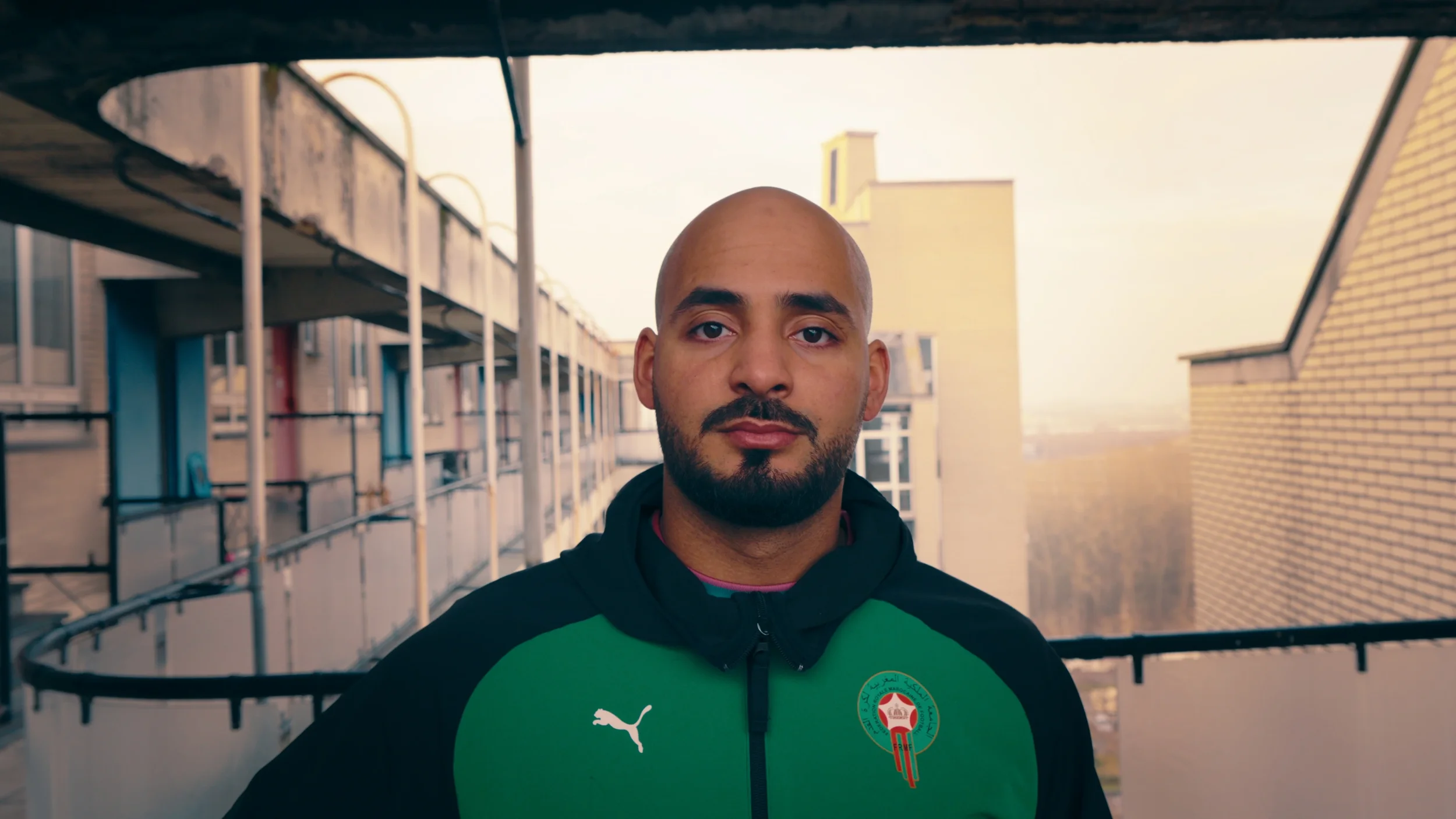 Man met een bald head en een baard, gekleed in een groen en zwart sportjack met het Puma en Algerije embleem, staat op een balkon met een stedelijke achtergrond.
