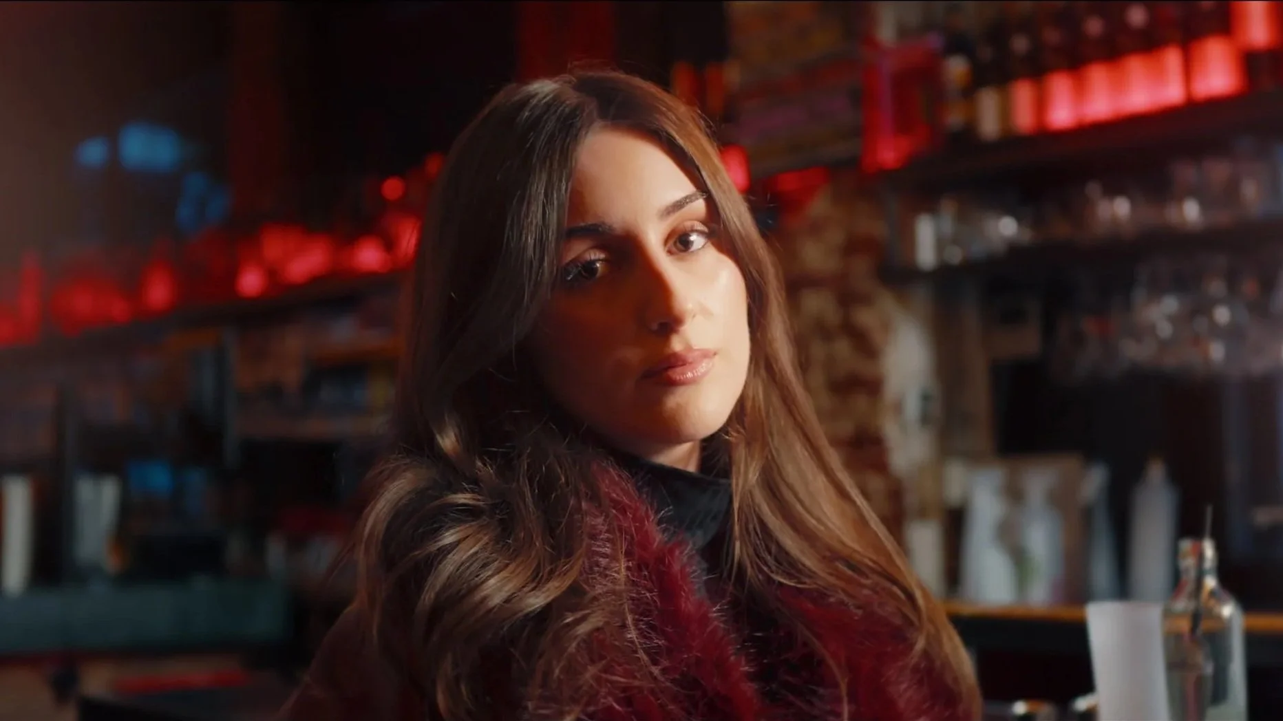 Een jonge vrouw met lang, golvend bruin haar, zittend in een bar of café, met een rustgevende uitdrukking.