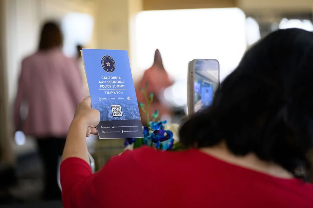 Person holding a blue pamphlet that reads 'California AAPI Economic Policy Summit, Thank You' and is taking a photo of it with a smartphone, at an indoor event.
