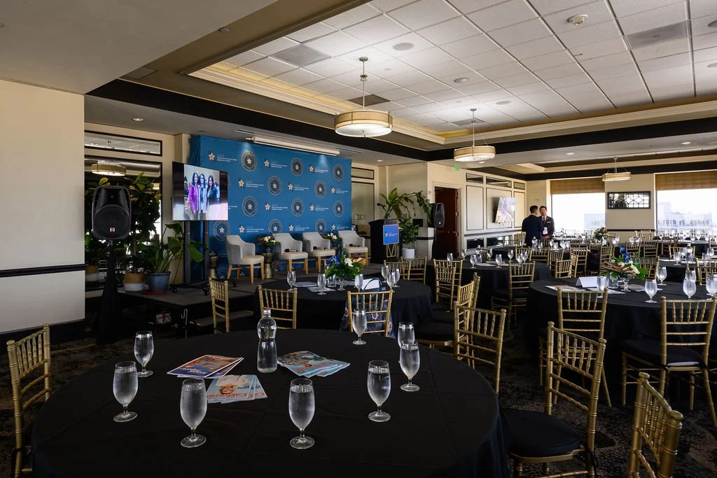 Event setup in a banquet room with round tables, black tablecloths, water glasses, and gold chairs. A stage with a blue backdrop, chairs, and TVs, with two people talking in front of the windowed wall.