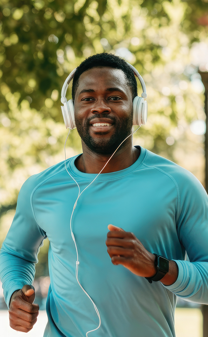 Man jogging outdoors wearing a blue long-sleeve athletic shirt, headphones, and a smartwatch, with trees and sunlight in the background.