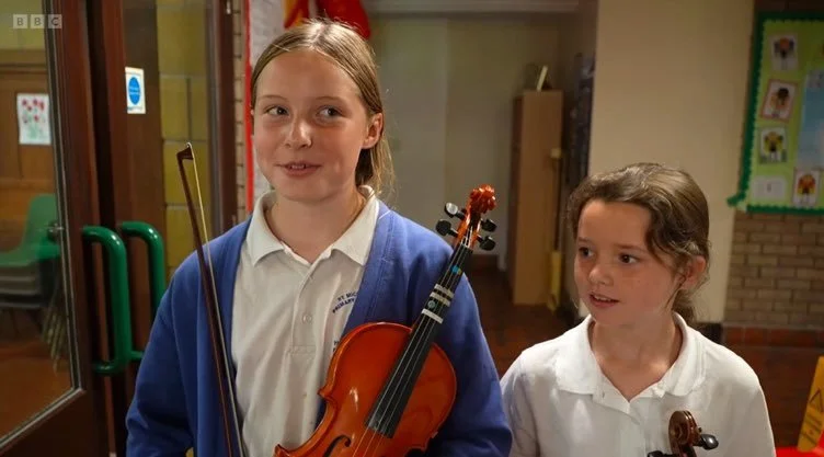 Two young girls in school uniforms holding violins inside a classroom.
