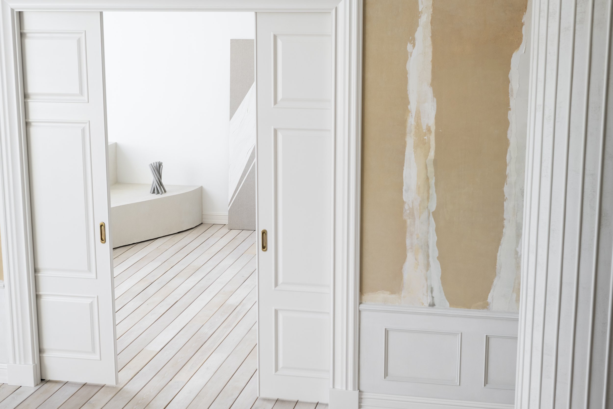 Intérieur minimaliste avec porte coulissante blanche et sol en bois clair, pièce adjacente avec mur inachevé et sculpture moderne. rénovation intérieurs appartement