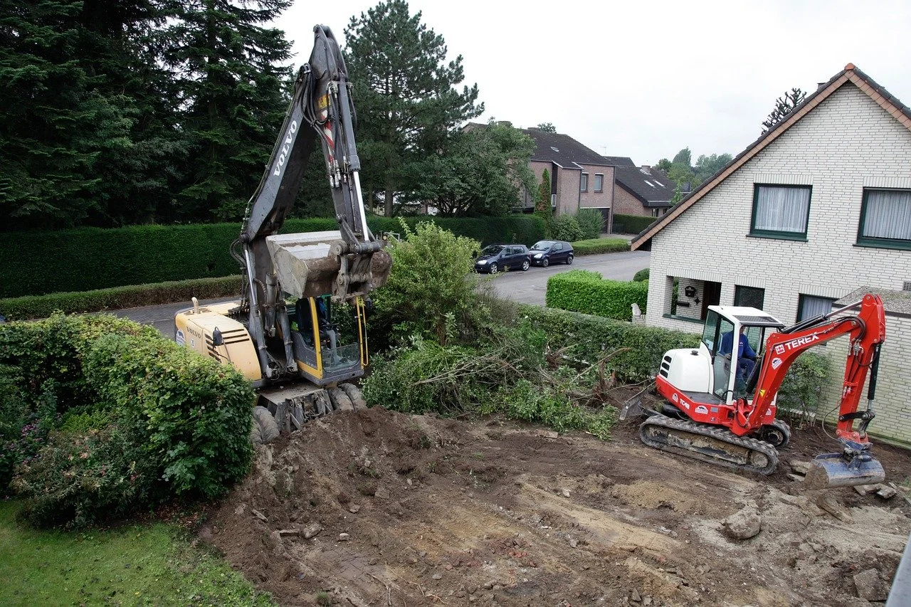 Deux pelles mécaniques creusant dans un jardin résidentiel, avec des arbustes et de la terre déplacée, près d'une maison en briques blanches.