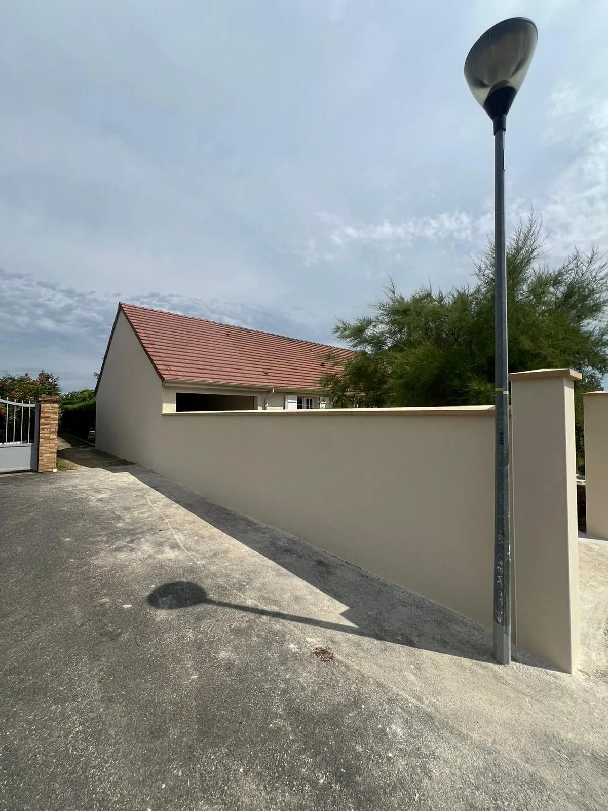 Allée avec un mur beige, un lampadaire et une maison au toit en tuiles, sous un ciel en partie nuageux. Construction de garage