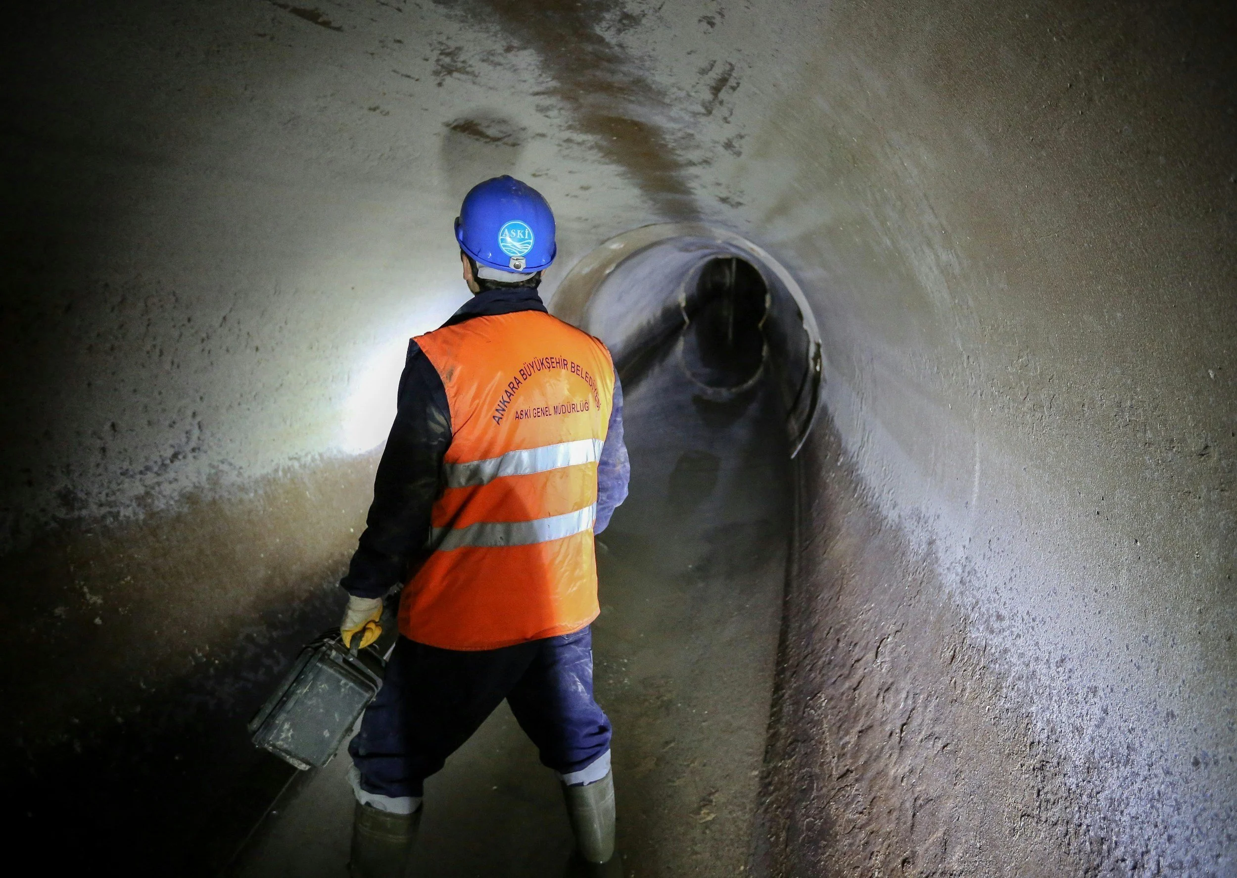 A worker in a blue helmet and orange safety vest standing in a tunnel, holding a toolbox, facing away from the camera and looking into the tunnel's dark interior.