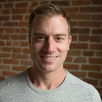 A man with short brown hair and a slight smile, wearing a light gray t-shirt, standing in front of a brick wall.