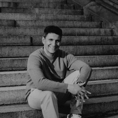 A young man sitting on a set of stairs, smiling at the camera, wearing a light-colored shirt and light pants.