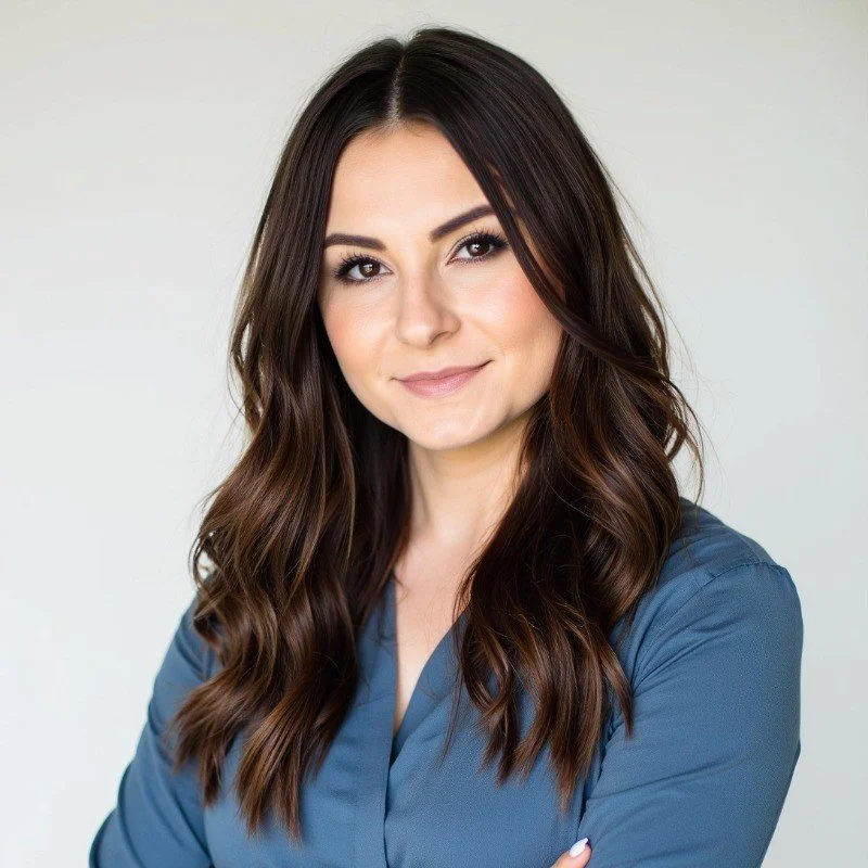 A young woman with long, wavy brown hair and light makeup, wearing a blue top, smiling softly with her arms crossed against a plain light background.