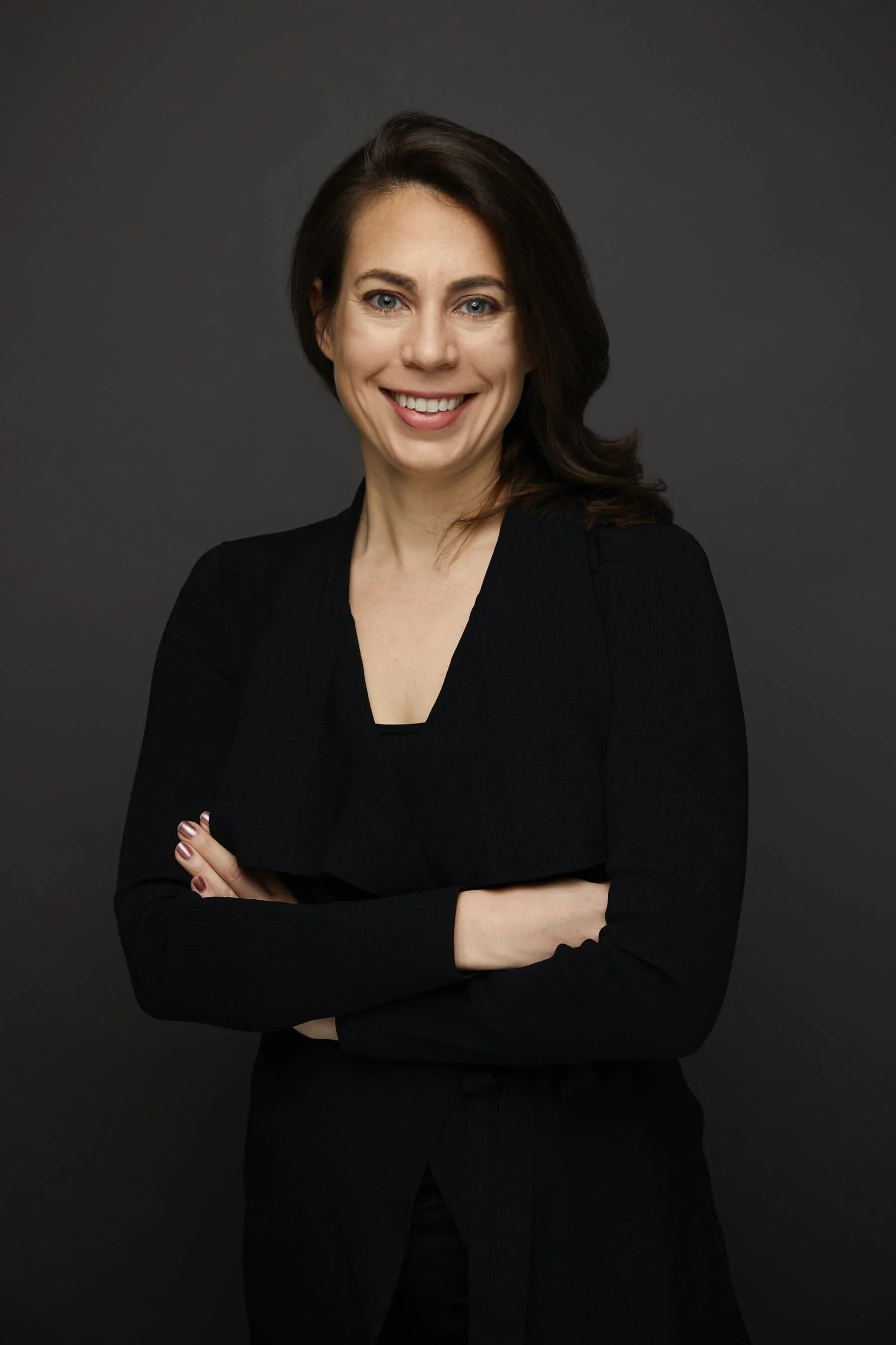 A woman with dark brown hair wearing a black blazer and black top, smiling with arms crossed, against a dark gray background.