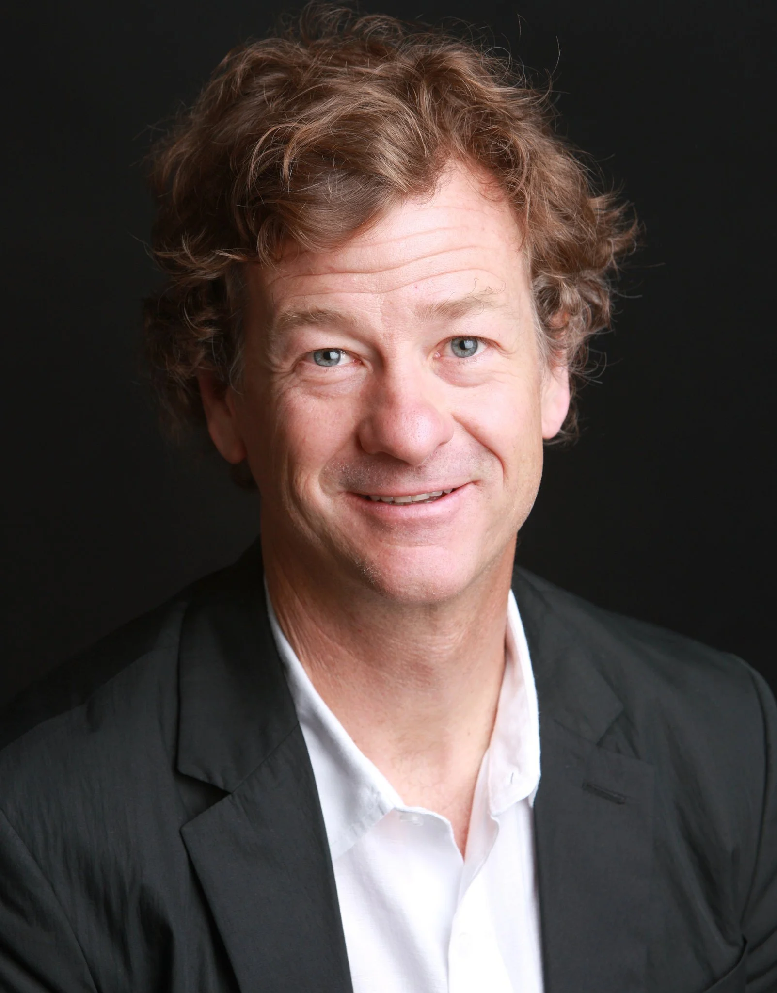 A portrait of a man with curly light brown hair, blue eyes, and a friendly smile, wearing a black blazer and white shirt against a black background.