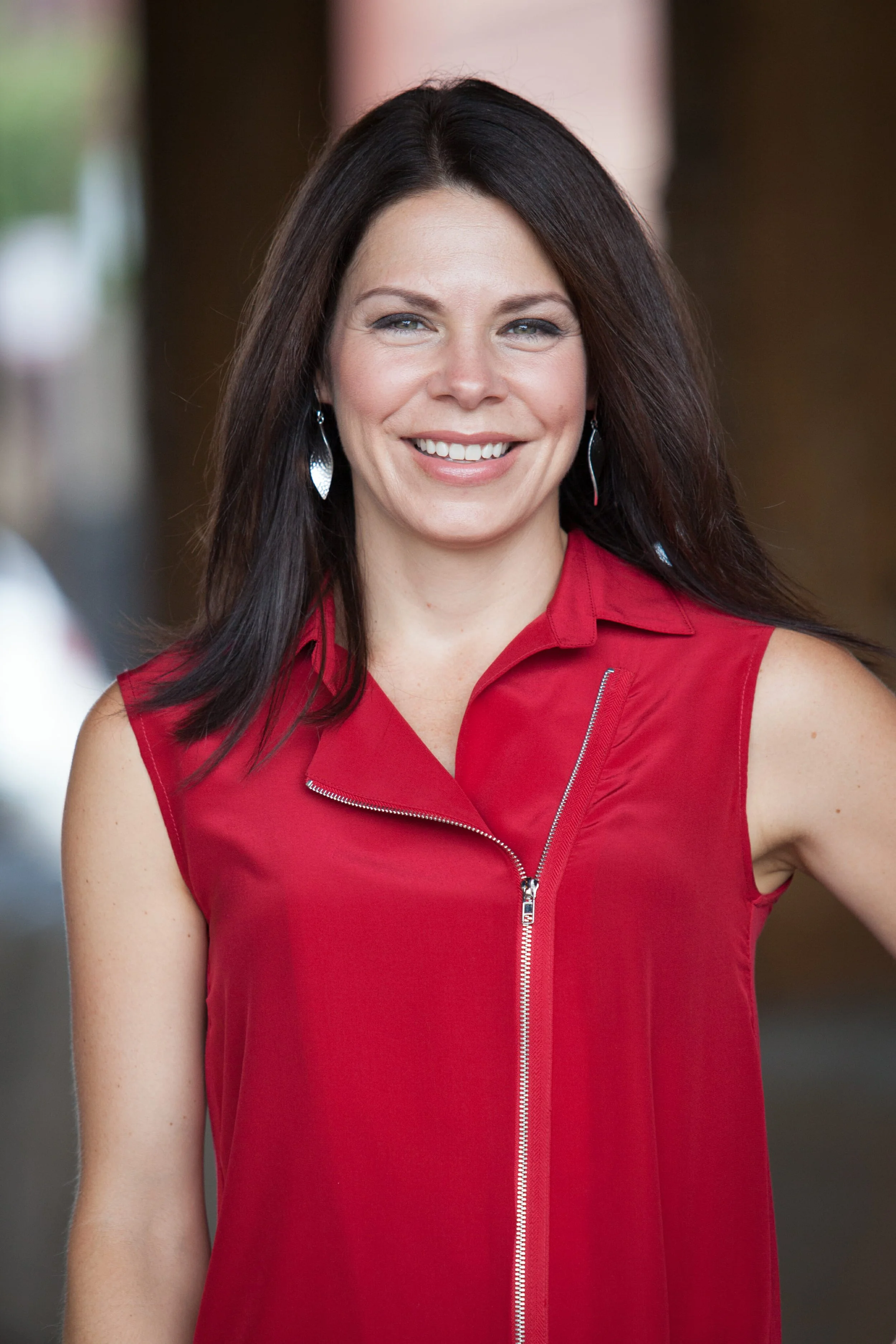 A woman with long dark hair, wearing a sleeveless red top with a front zipper, smiling outdoors.
