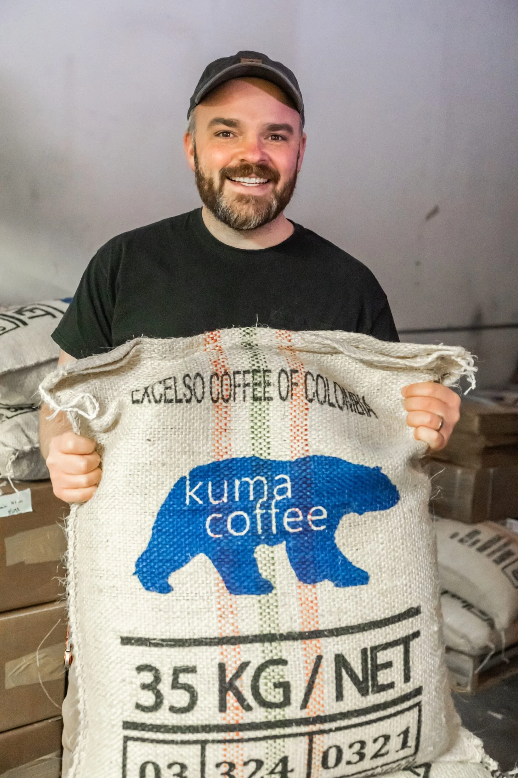 A man with a beard and cap smiling while holding a large bag of coffee beans labeled 'Kuma Coffee,' '35 KG NET.'