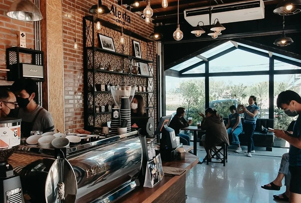 Young coffee business owners inside a cozy coffee shop with large windows, brick walls, and modern hanging lights, enjoying their drinks and working on laptops.