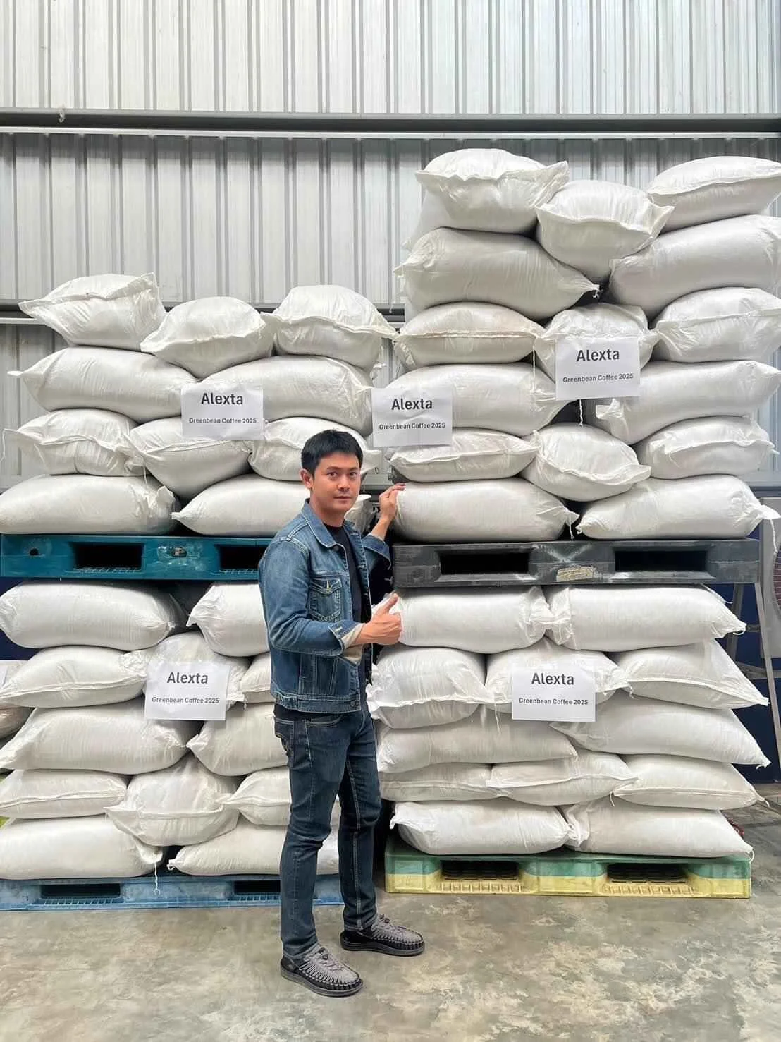 Alexta in a denim jacket and jeans standing in front of large stacked bags of coffee beans labeled 'Alexta' and 'Greenbean Coffee 2025' in a warehouse setting.