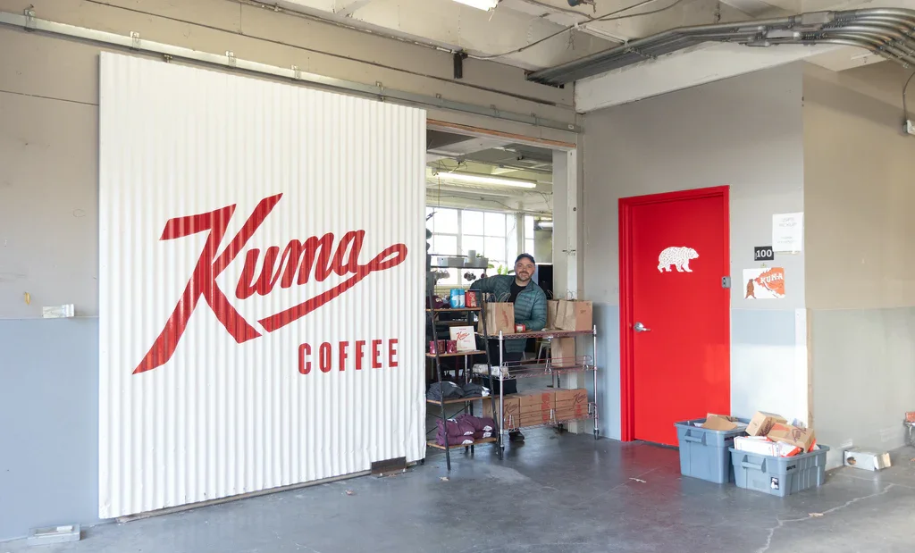 Interior of a coffee shop with a large sign that reads 'Kuma Coffee'. A man stands behind a cart filled with boxes, and there is a red door with a white bear silhouette on it. Two gray bins with packages are on the floor near the door.