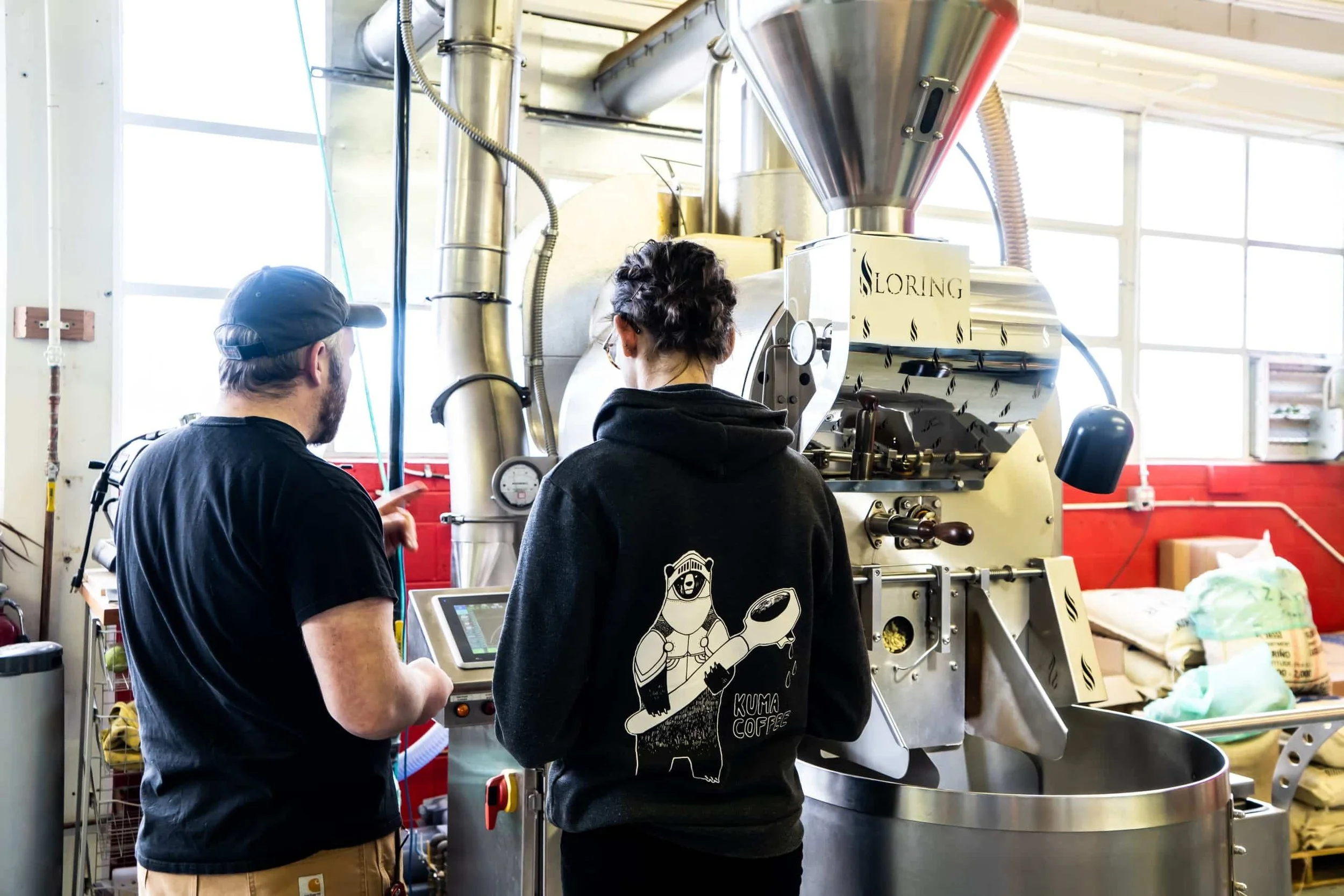 Two people are operating a large coffee roasting machine in a brightly lit industrial setting. One person is pointing at the machine, while the other is managing the controls. The person on the right is wearing a black hoodie with a bear character and the words 'Kuma Coffee' on the back.