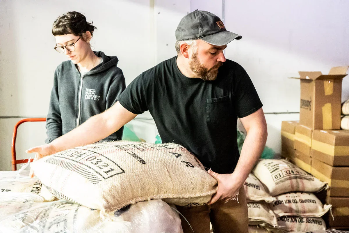 A man wearing a gray cap and black shirt stacking a large burlap bag labeled "Luzern" in a warehouse. A woman with glasses and dark hair in a gray hoodie with "MUMA COFFEE" stands nearby, surrounded by cardboard boxes and more burlap bags.