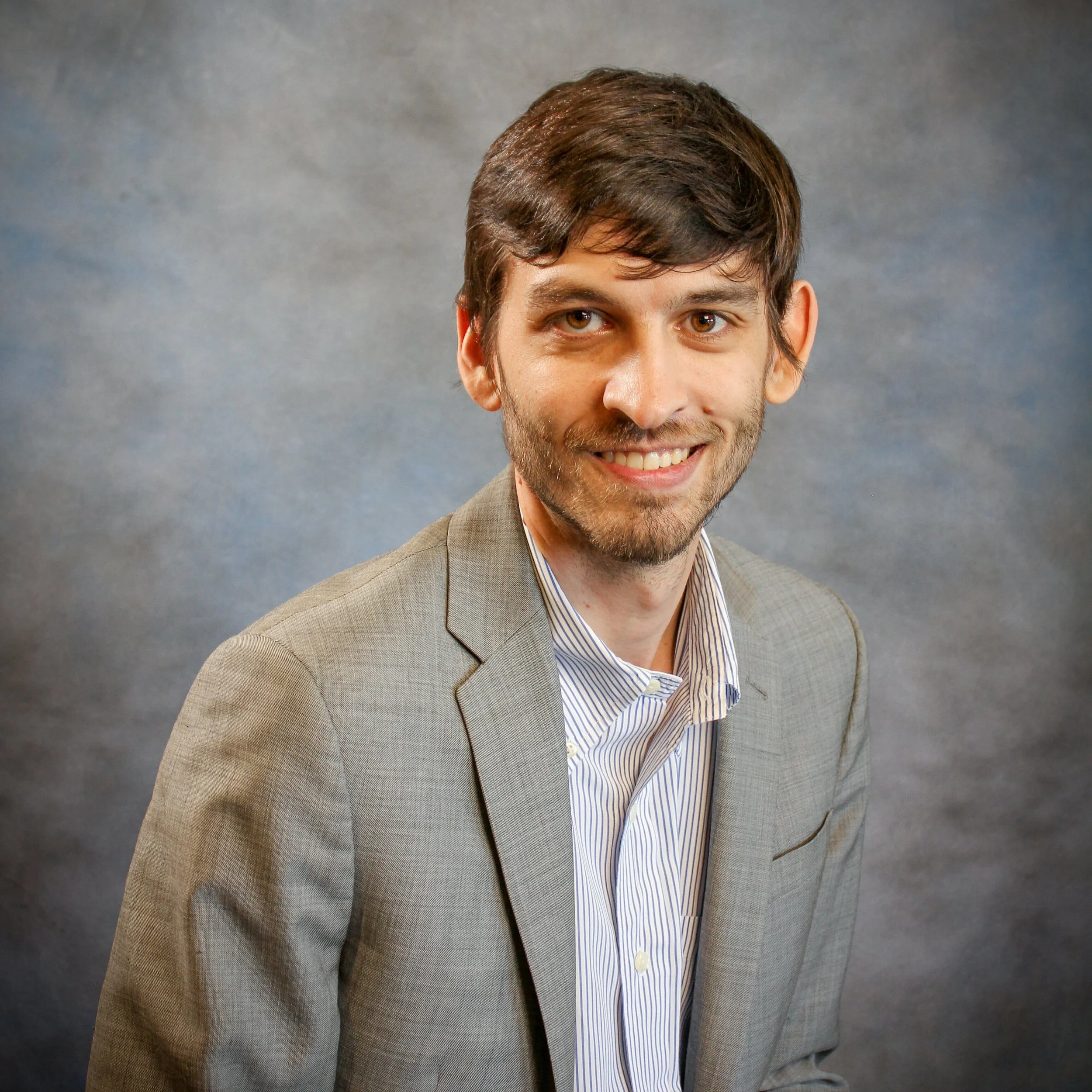 A smiling man with dark brown hair, a beard, and dressed in a gray suit and striped shirt poses against a gray background.
