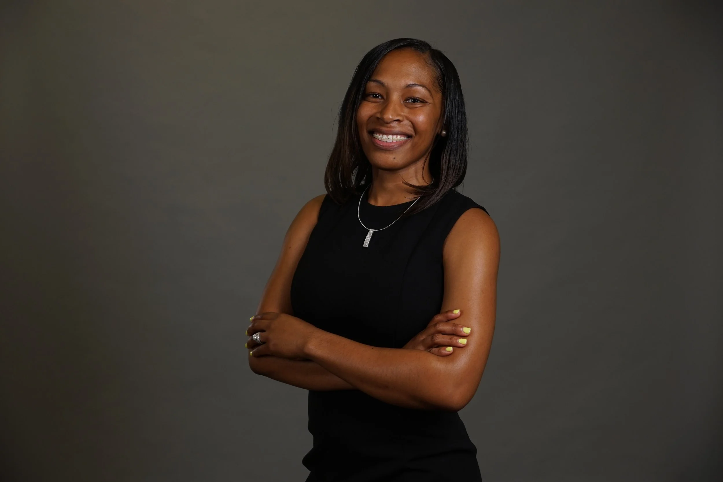 A confident woman with shoulder-length black hair, wearing a sleeveless black dress, standing with her arms crossed and smiling against a dark grey background.