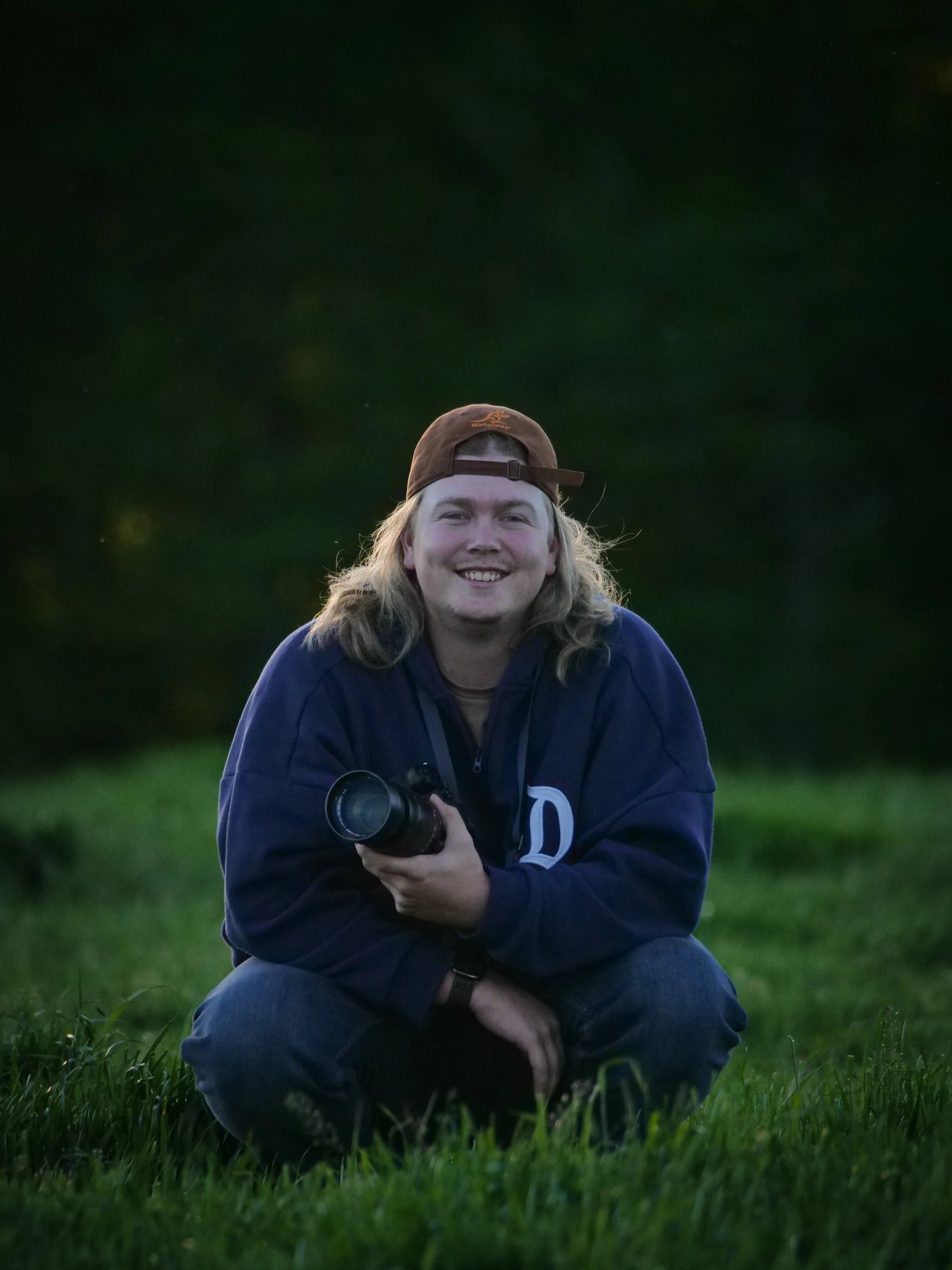 Person sitting on grass holding a camera, wearing a cap and a dark sweatshirt, with a blurred green background.
