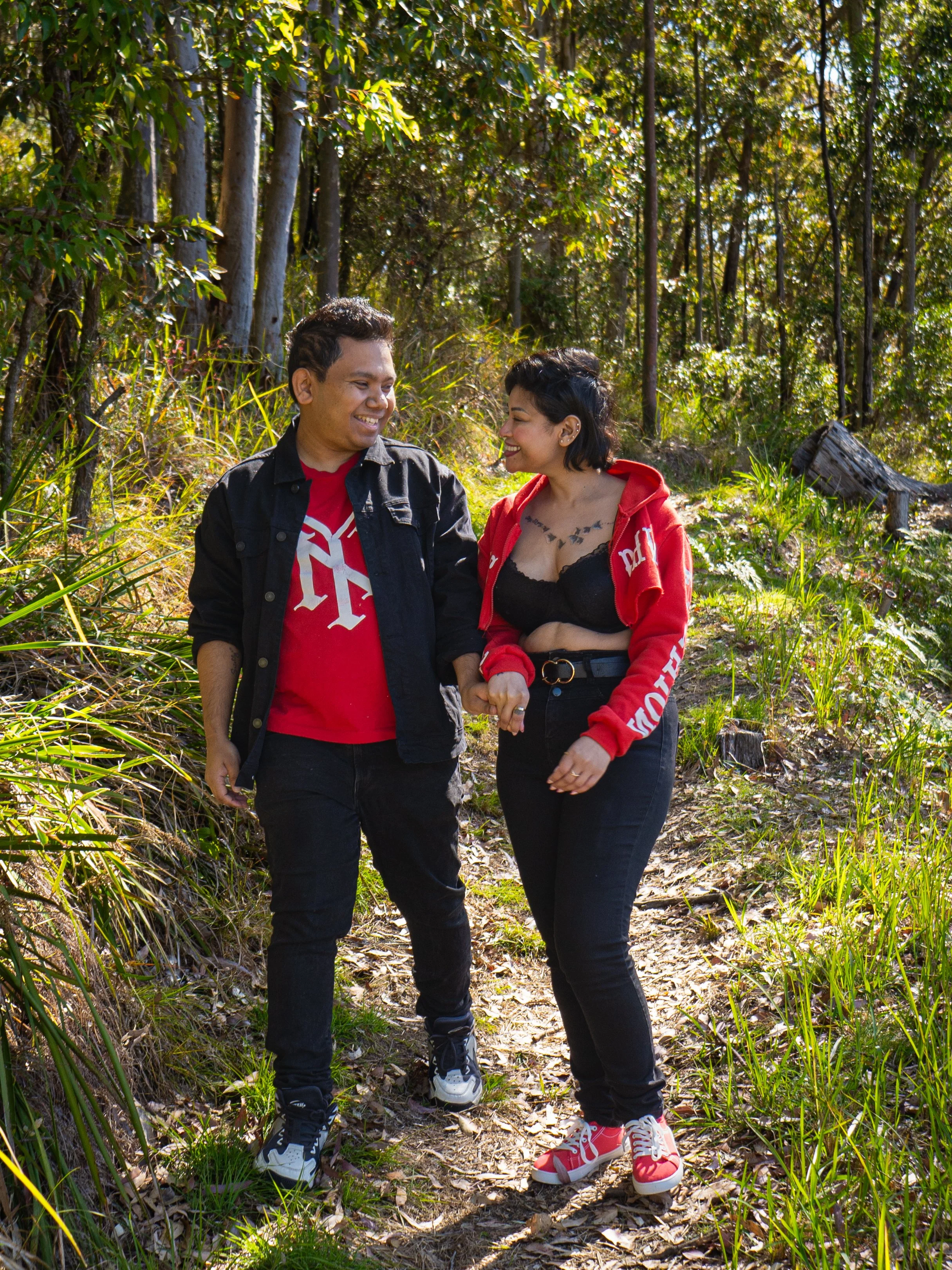 A couple walking through a wooded area, smiling at each other. The man is wearing a black jacket and red shirt, and the woman is wearing a red hoodie and black pants. Sunlight filters through the trees, illuminating the greenery around them.