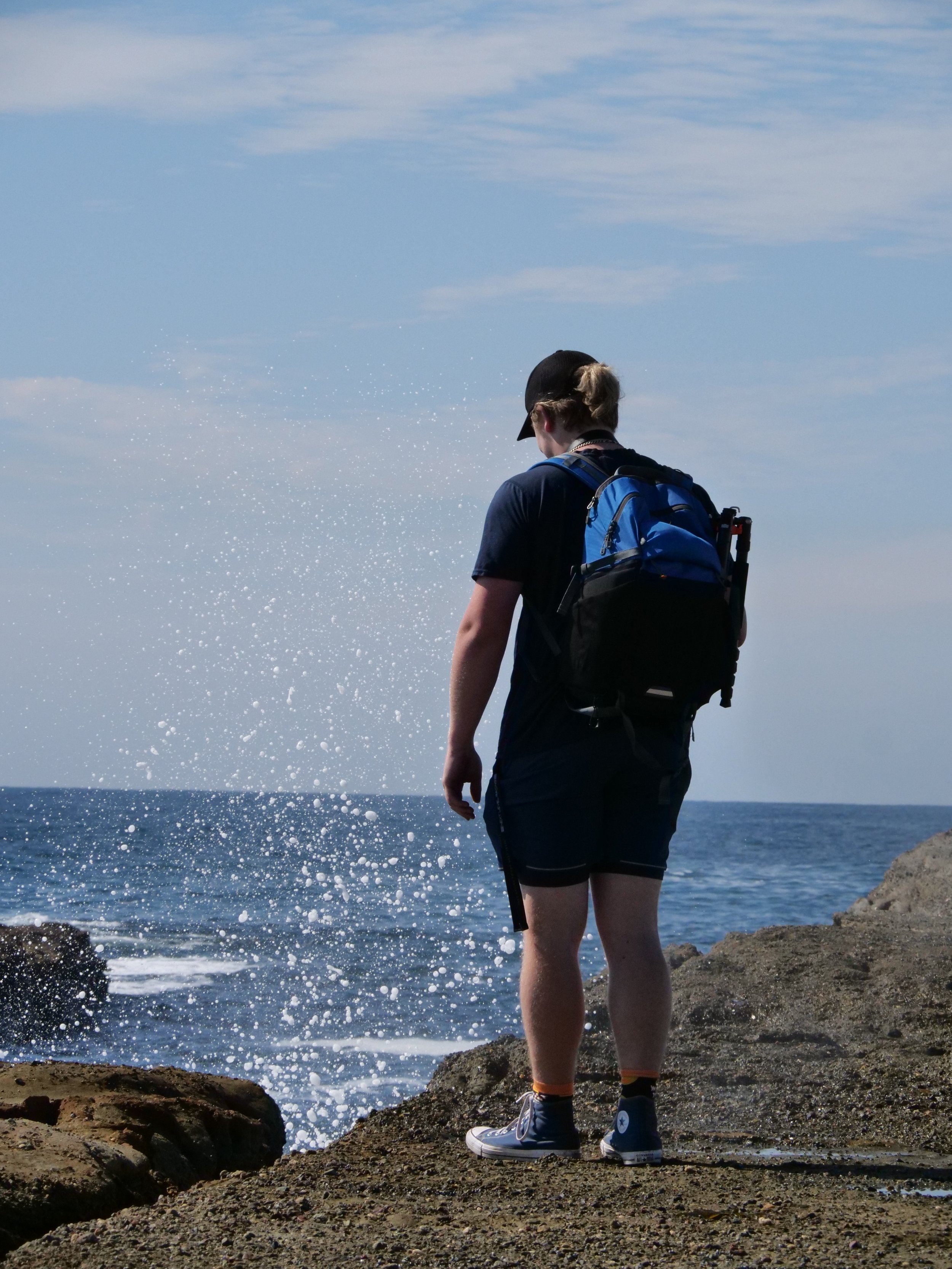 A person wearing a backpack stands on a rocky shoreline, observing ocean waves under a blue sky.