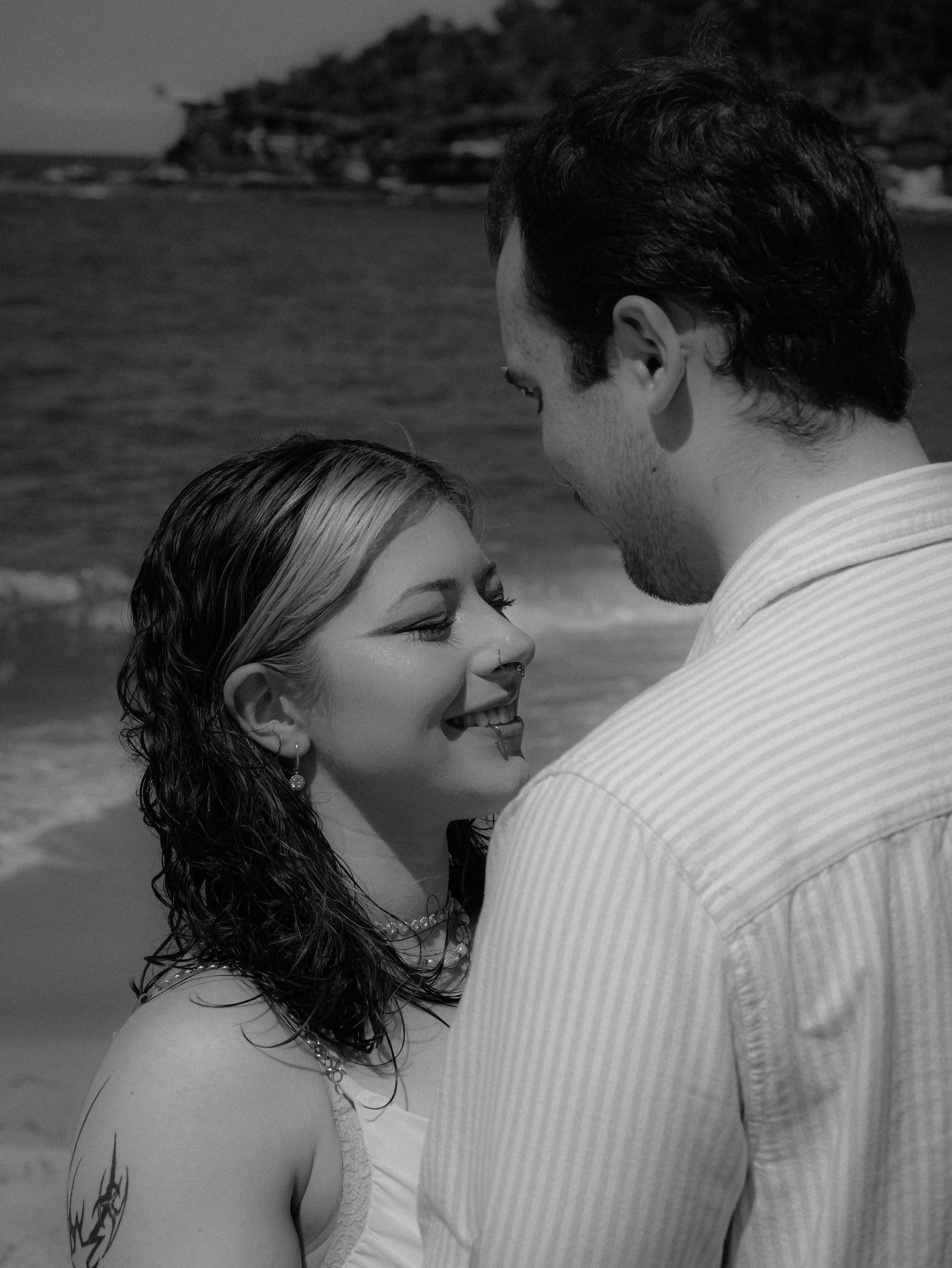A couple smiling at each other on a beach, with the ocean and rocky shoreline in the background. The woman has wet hair and a tattoo on her arm, while the man wears a striped shirt.
