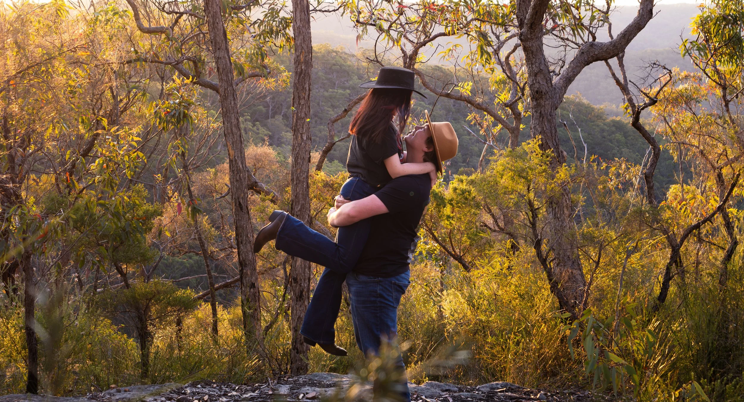 A couple embracing in a forest, with one person lifting the other. Both are wearing hats and jeans. The sun is setting, casting a warm glow on the trees around them.