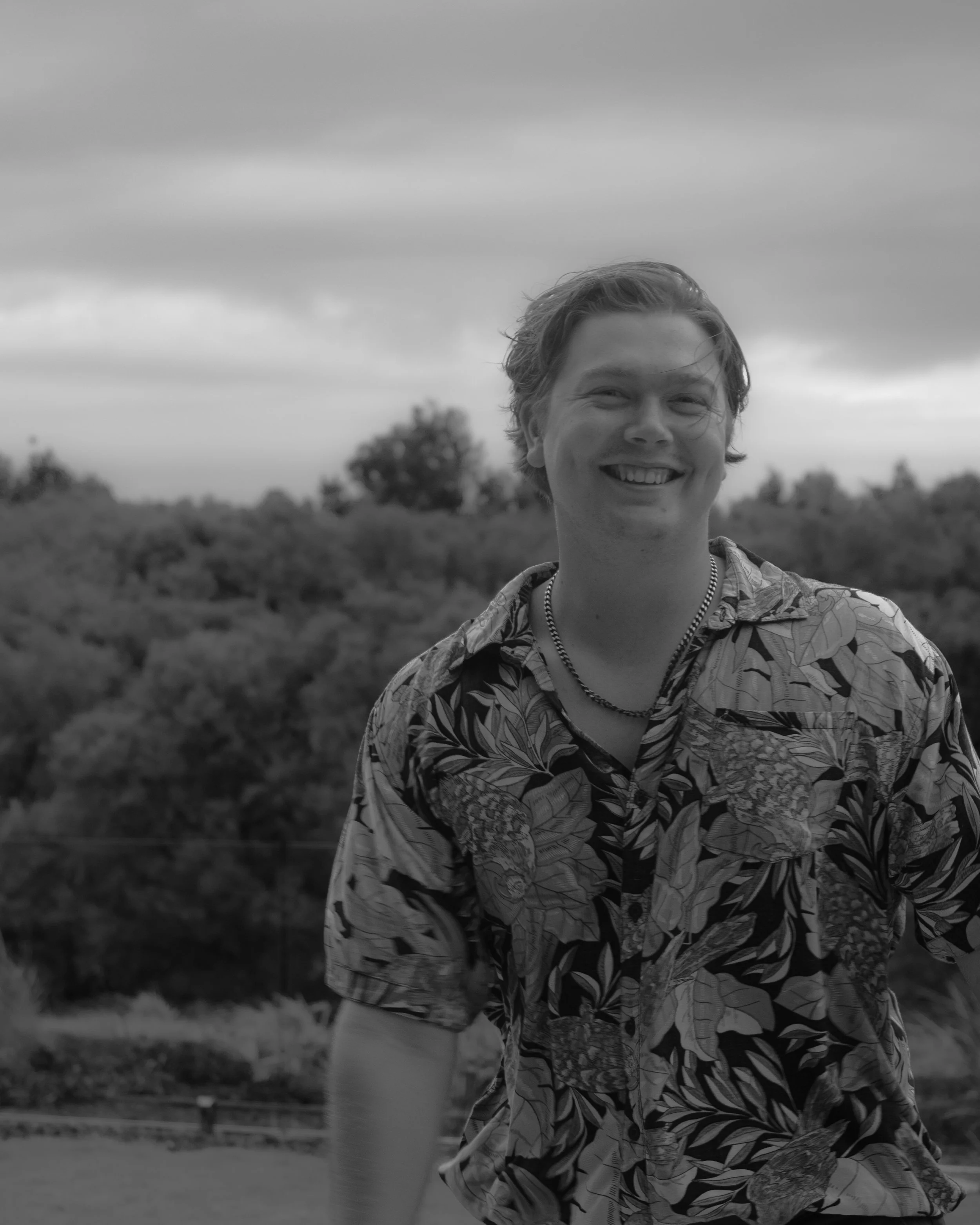 Black and white photo of a smiling person wearing a floral shirt, standing outdoors with trees and a cloudy sky in the background.