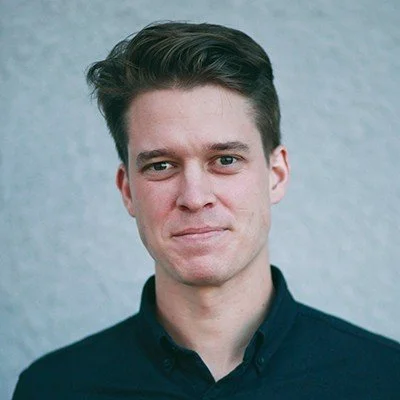 A man with short brown hair wearing a dark button-up shirt, standing against a textured wall.
