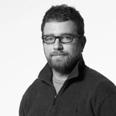 Black and white photo of a man with short hair, wearing glasses and a zip-up sweater, looking at the camera.