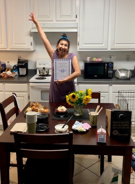 A woman standing in a kitchen, smiling and raising one arm, holding a framed diploma or certificate. The kitchen counter has a microwave, toaster, and other appliances. A dining table in front has a bouquet of sunflowers, cupcakes, snacks, disposable