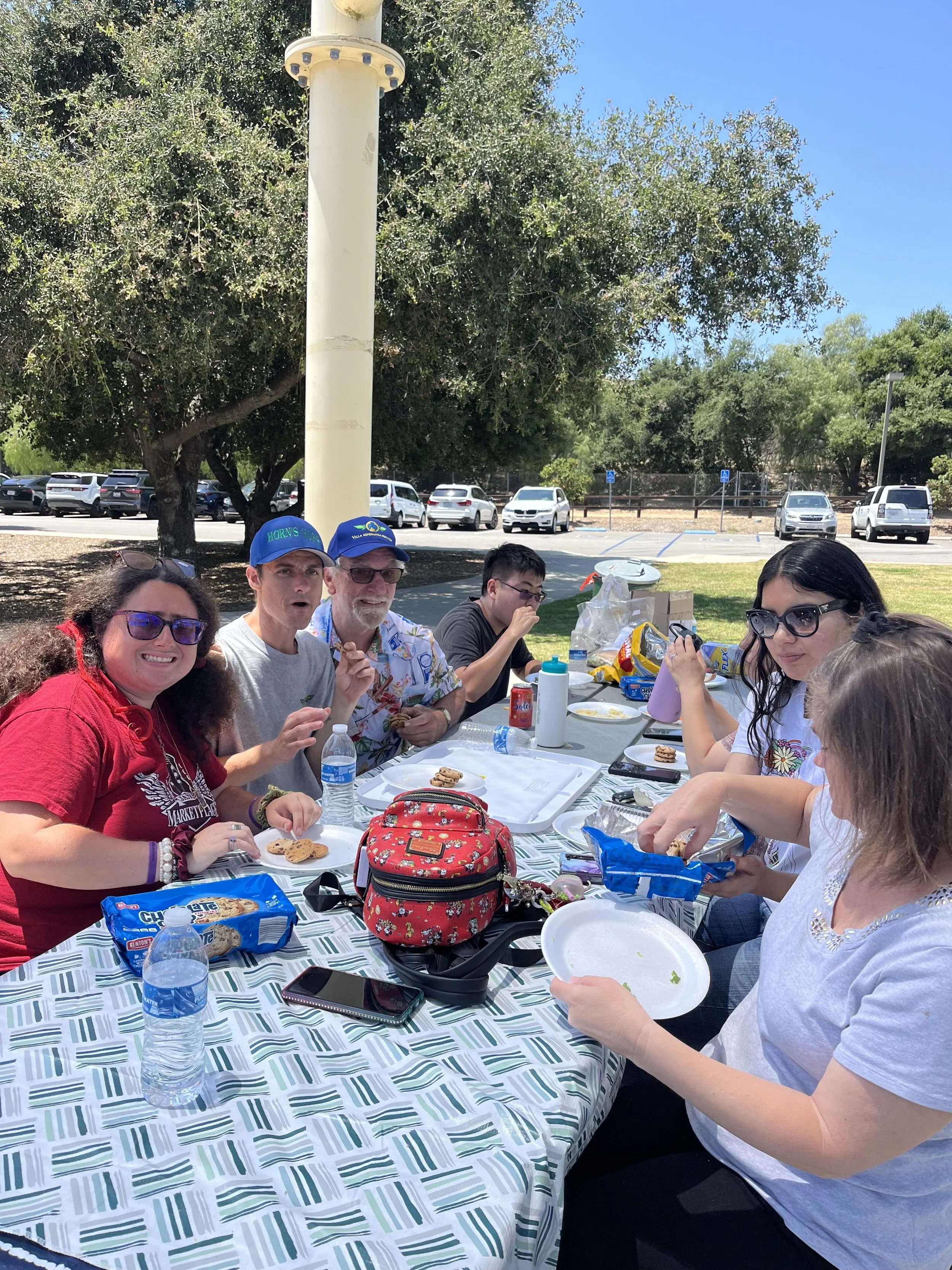 Group of people sitting at an outdoor picnic table enjoying snacks on a sunny day.
