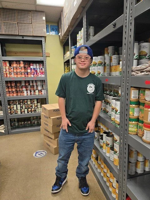 Young man standing in a grocery store aisle, wearing a green T-shirt, jeans, and a blue backwards cap, surrounded by shelves of food products.