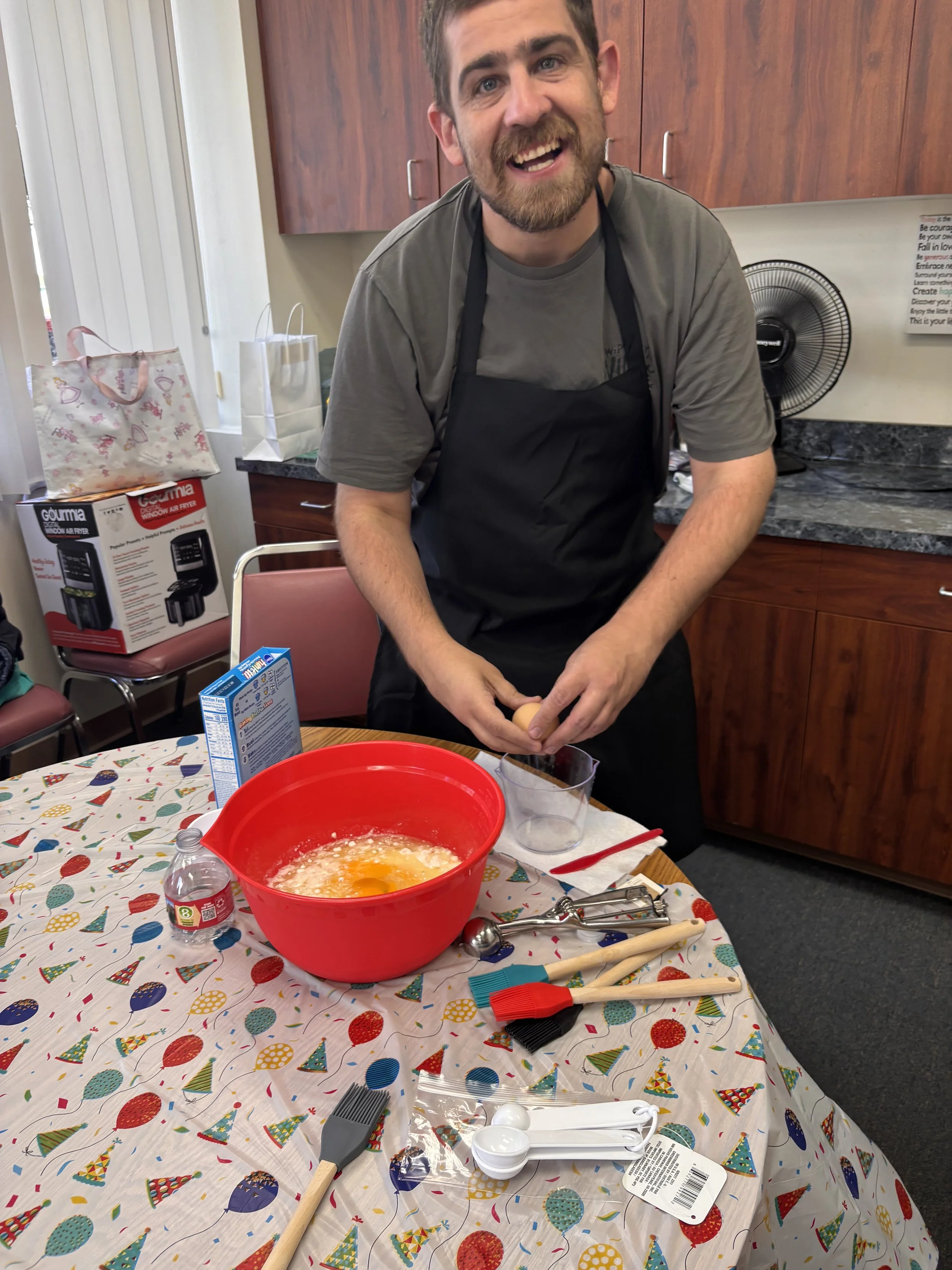 Man with beard and short hair cracking an egg into a glass bowl in a kitchen, smiling at the camera, surrounded by baking tools and ingredients on a table with a holiday-themed tablecloth.