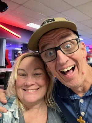 Two smiling people taking a selfie in an arcade, with colorful neon lights in the background.