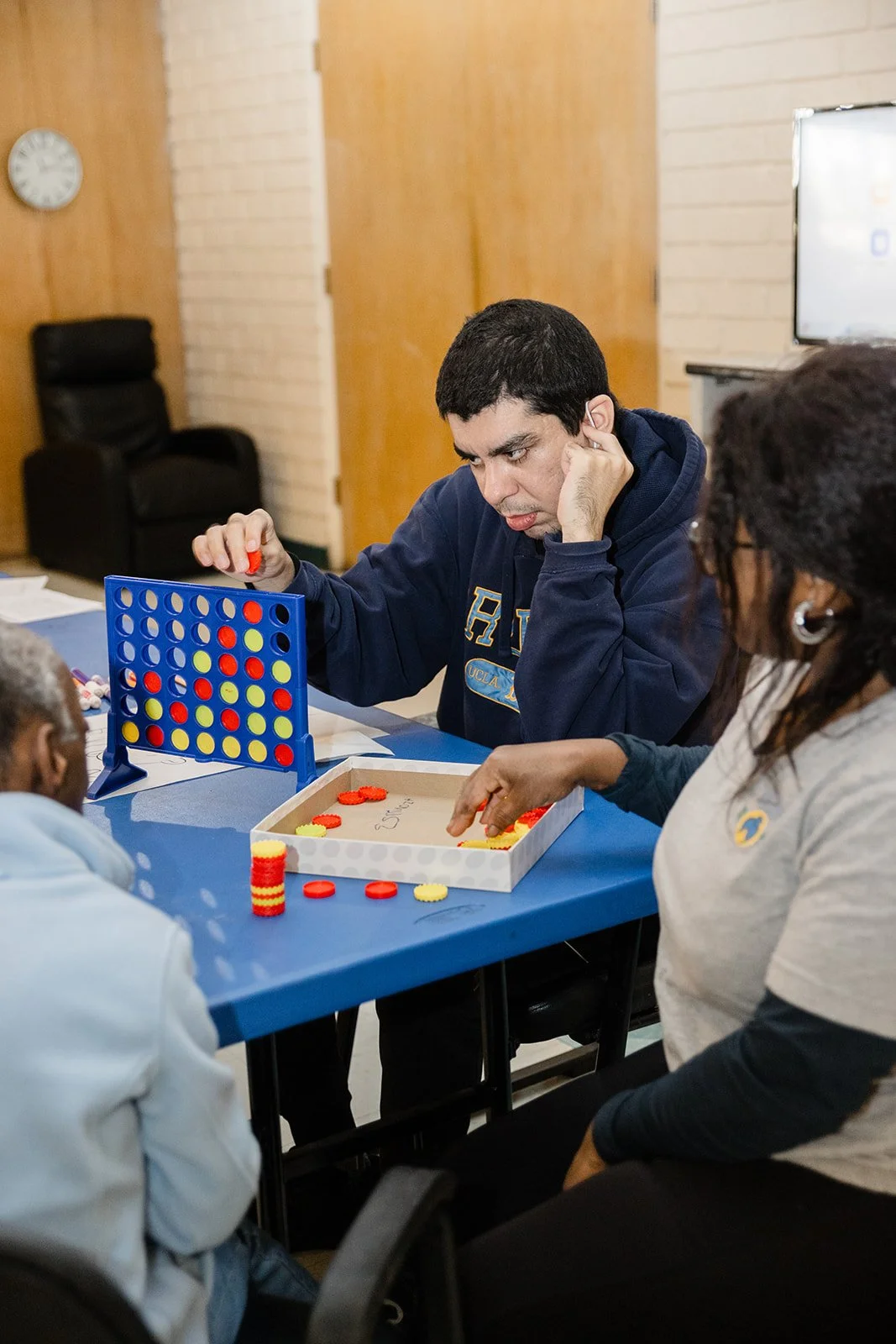 People playing a game of Connect Four at a blue table, with a female support aide handing game pieces to a male participant, in a room with wooden walls and a clock on the wall.