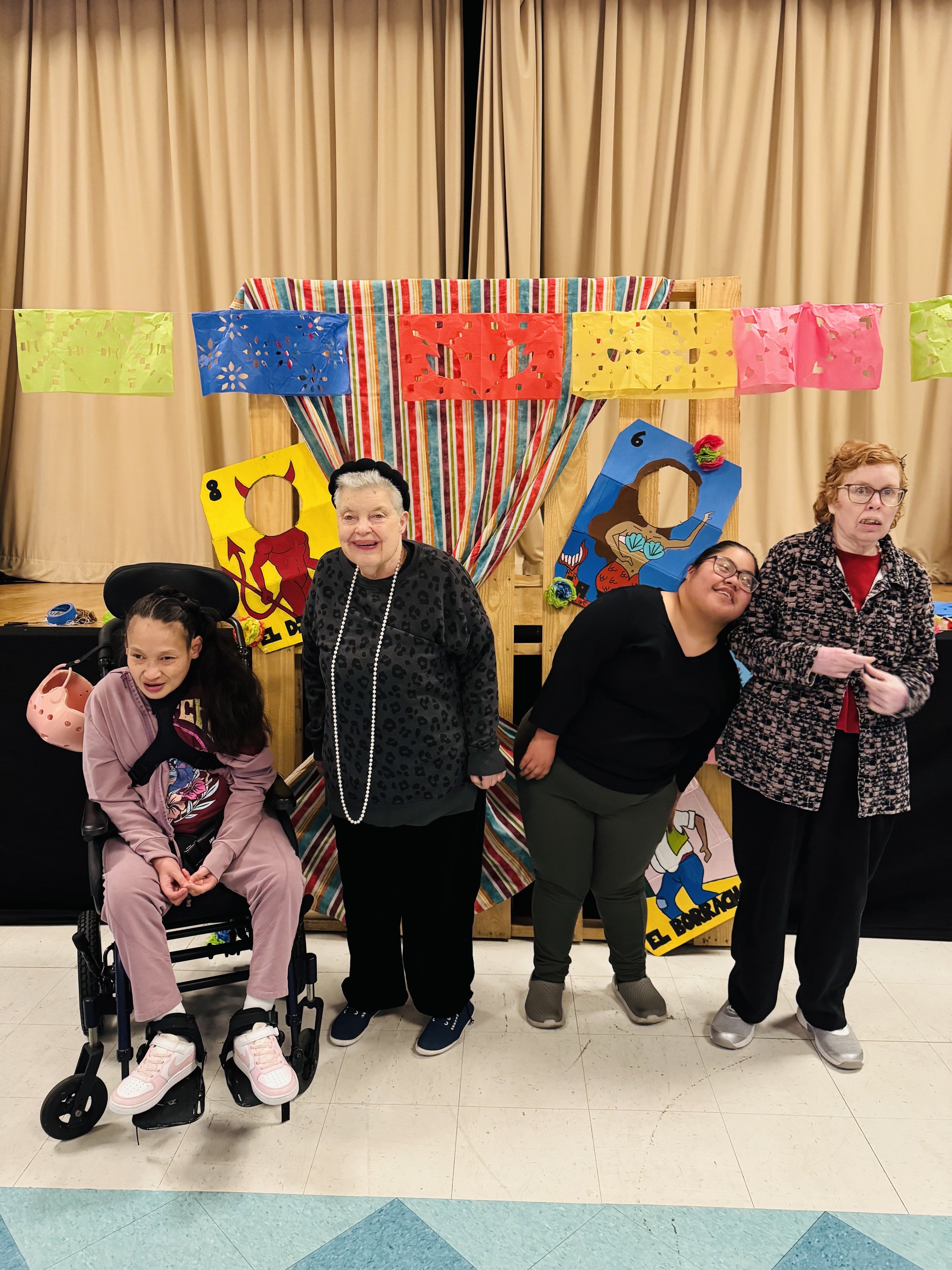 Four people standing in front of a colorful puppet show backdrop with banners. One person is in a wheelchair, smiling, with a helmet hanging on their chair. The other three women are standing next to her, smiling.