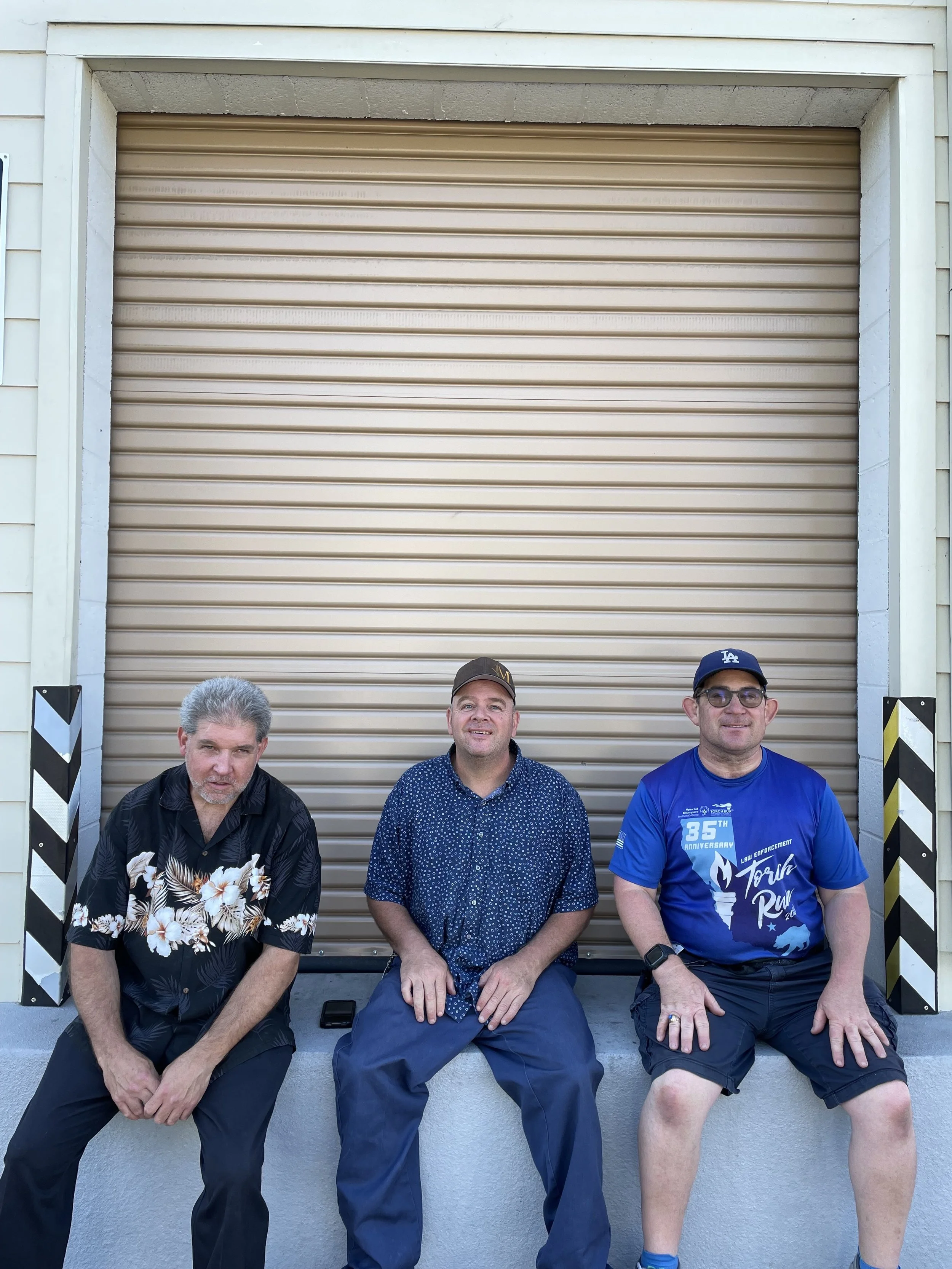Three men sitting on a concrete ledge in front of a beige garage door, with black and white striped safety barriers on each side.