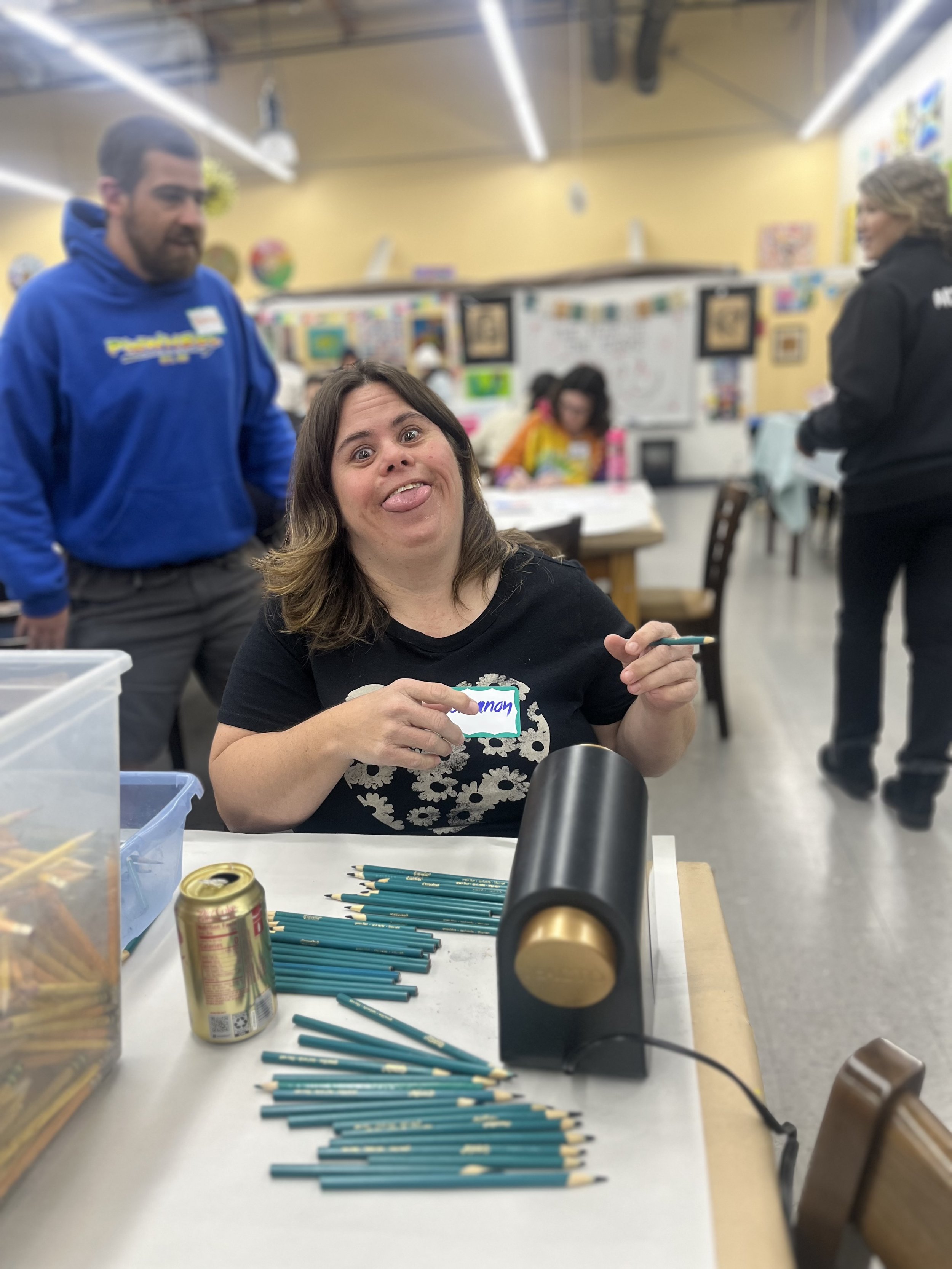 A woman sitting at a table with scattered teal-colored pencils, holding a small item, and making a funny face. In the background, there are people and colorful decorations in a classroom or community center setting.