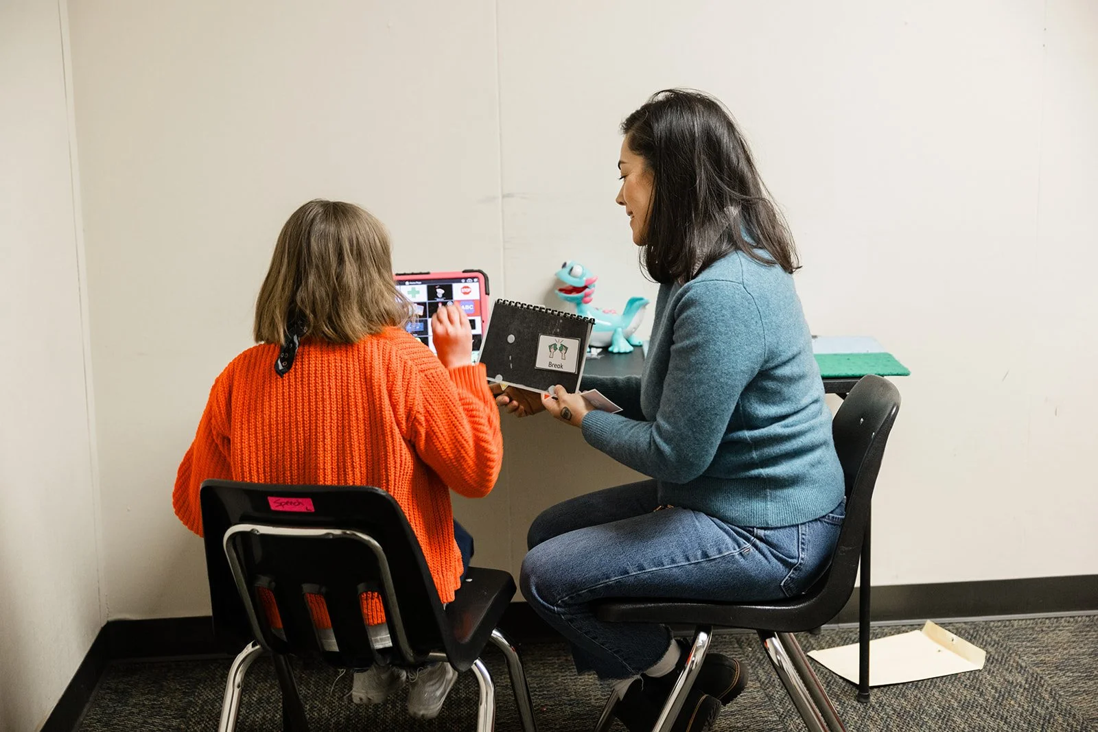 A young girl and a woman sitting in a room with white walls, looking at a tablet together. The girl is wearing an orange sweater, and the woman is wearing a teal sweater and jeans. They are engaging in speech therapy.