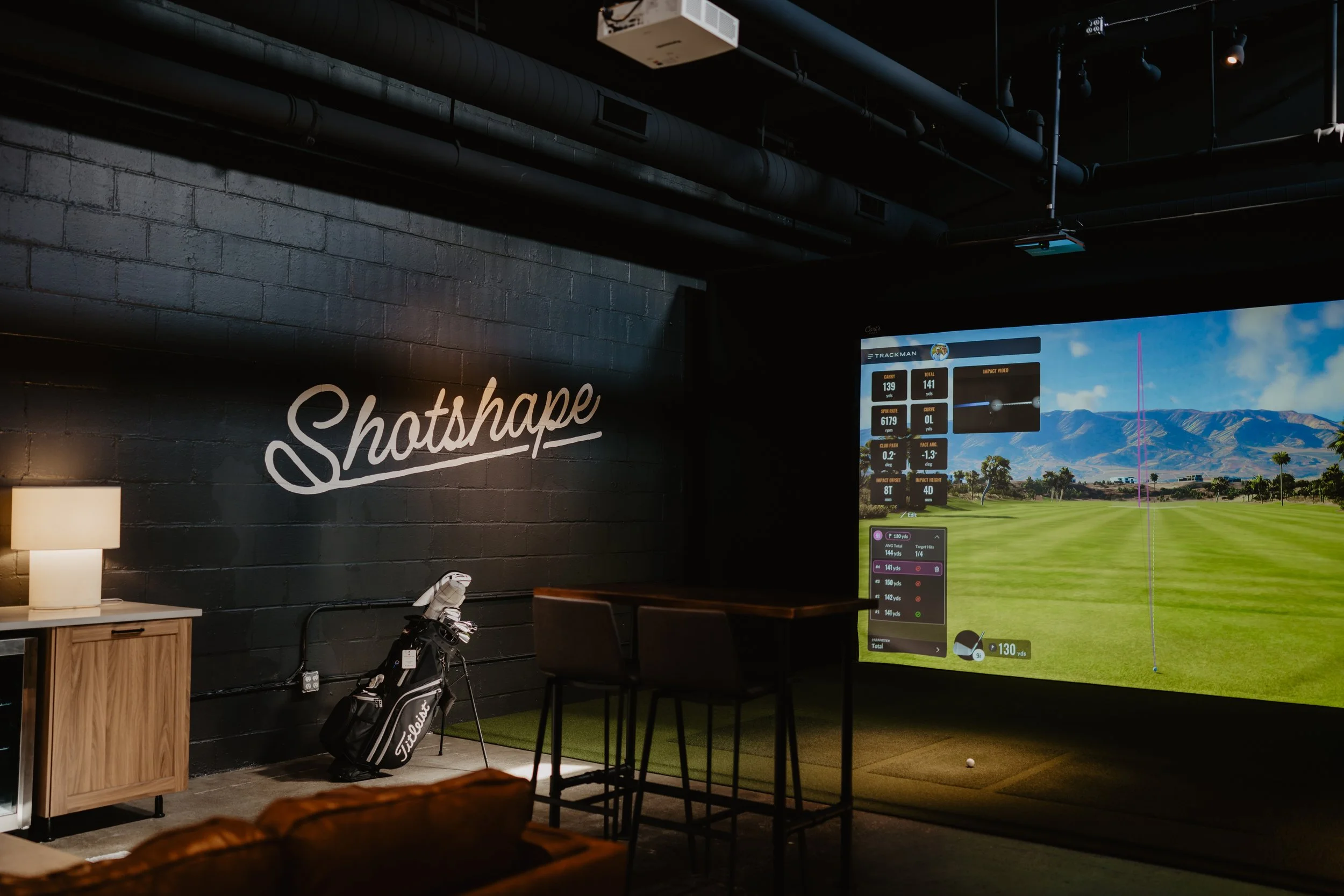 Indoor golf simulation room with a large screen displaying a golf course, a golf bag with clubs, a small wooden cabinet with a lamp, and a high table with chairs. The wall has the word 'Shotshape' written in cursive.