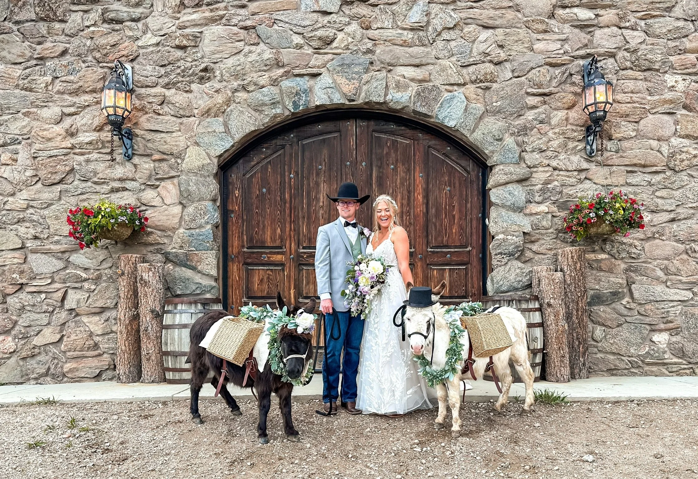 A bride and groom standing in front of a wooden door at a rustic venue, surrounded by stone walls, with two decorated donkeys at their sides.
