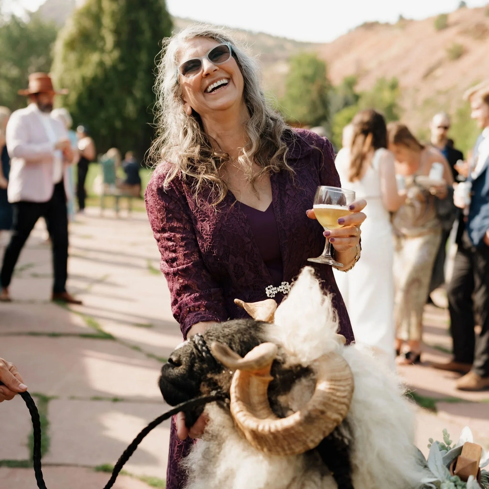 A woman with gray hair and sunglasses smiling at an outdoor event, holding a glass of white wine, with a ram costume in front of her, and people in the background.