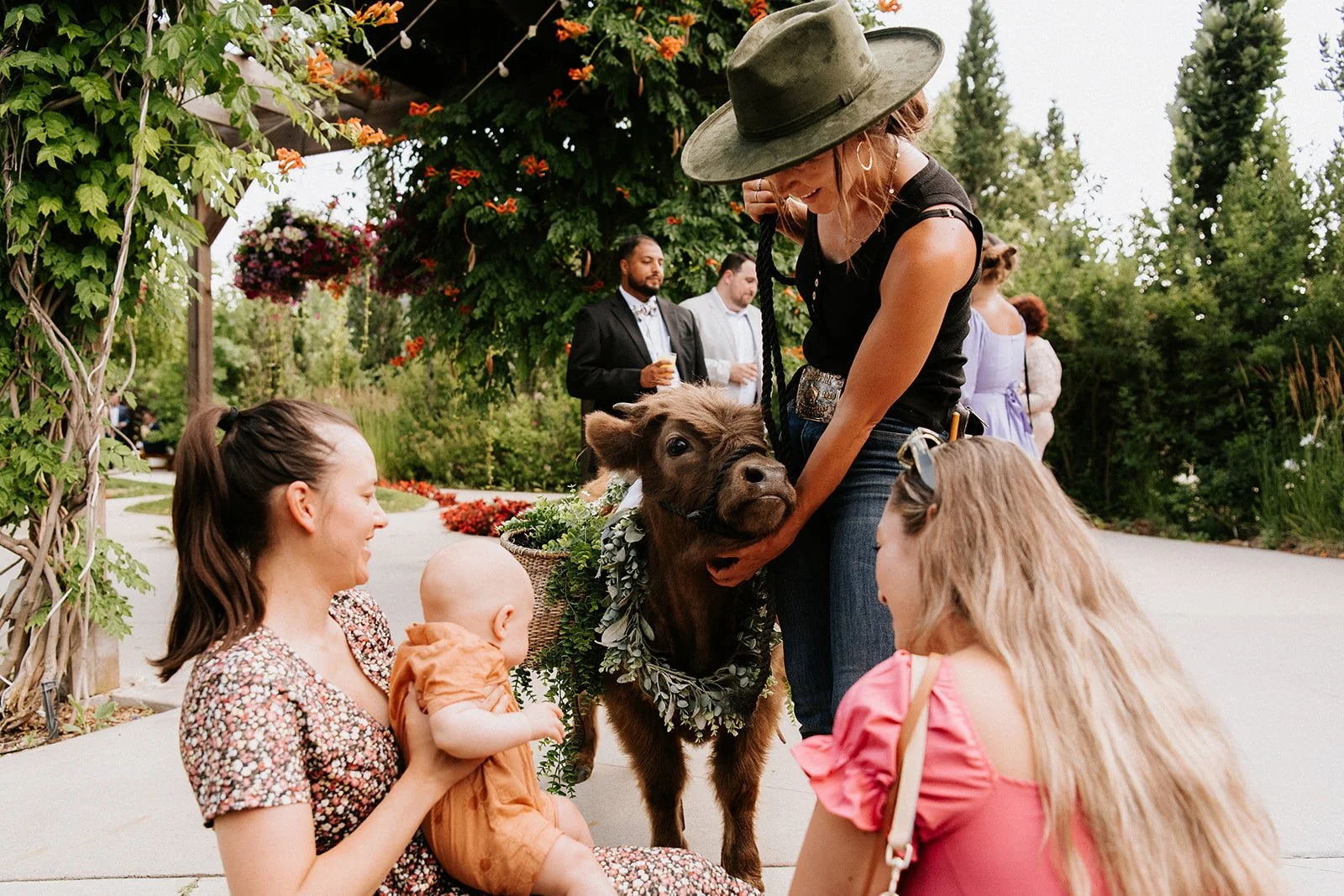 People at an outdoor event interacting with a small pig. A woman in a black top and hat is holding the pig's face, and a woman with long blonde hair and a pink dress is kneeling nearby. A woman with a floral dress and a young child are sitting on the ground looking at the pig. Other people are in the background, some holding drinks, with lush green trees and flowers surrounding the scene.