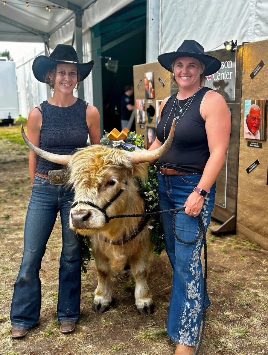 Two women wearing cowboy hats, standing on either side of a decorated cow with large horns, in an outdoor setting under a tent, with display boards behind them. The women are smiling and holding the cow's lead.