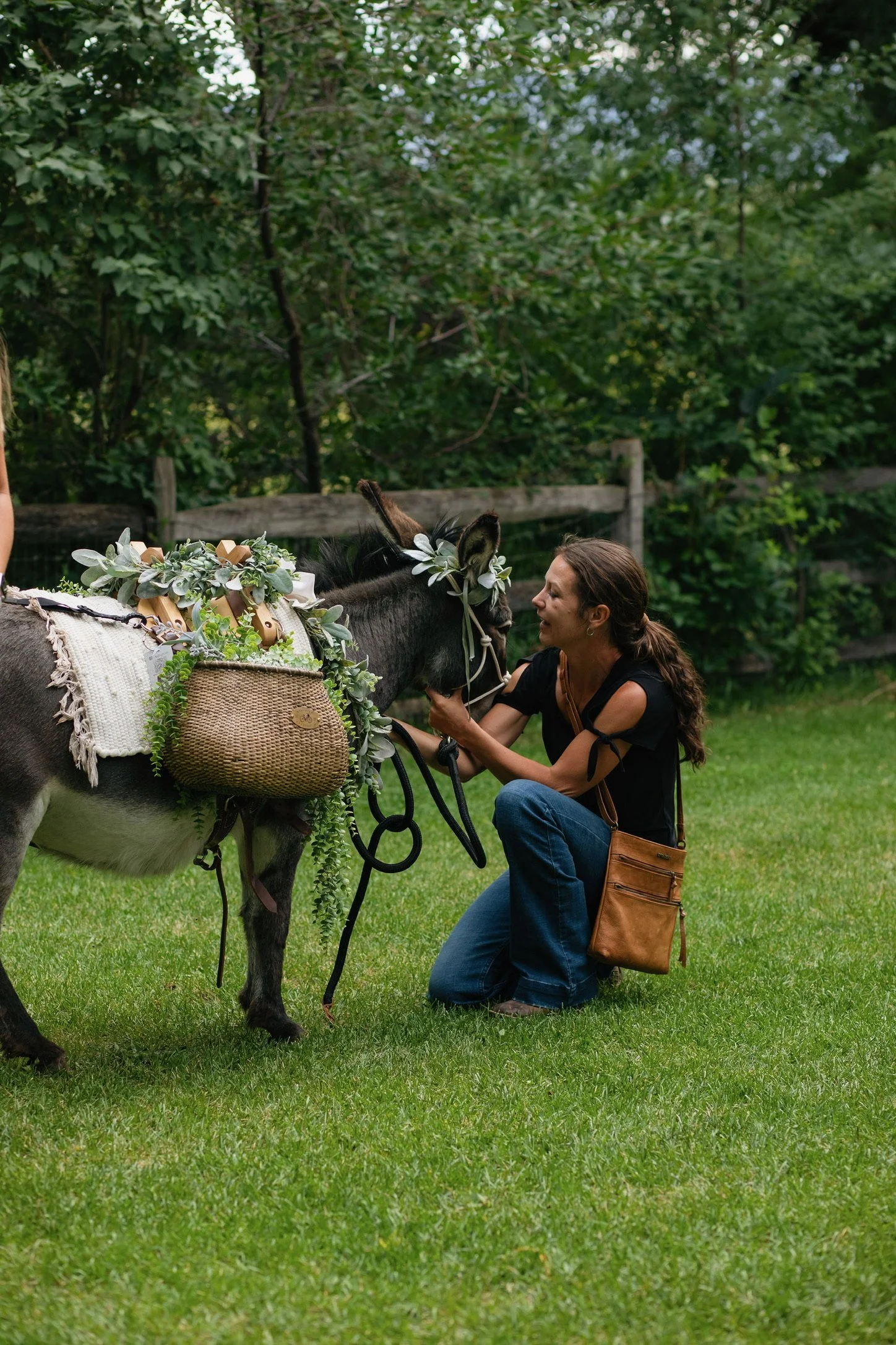 A woman kneeling on the grass and smiling at a donkey decorated with flowers and greenery in an outdoor park or garden.